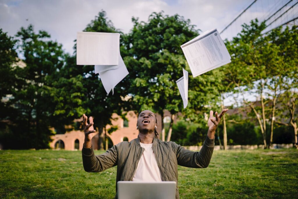 Young student happy with GCSE results, throwing papers in the air outdoors with laptop on lap